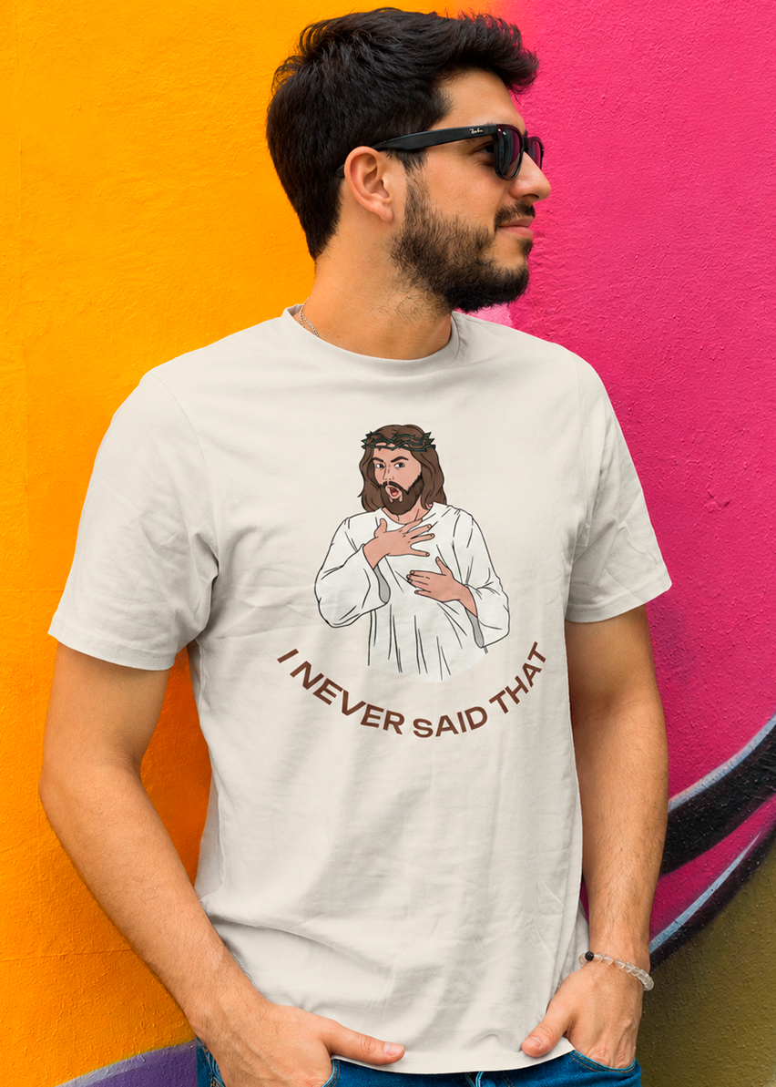 A man wearing a light gray graphic t-shirt with a cartoon illustration of Jesus and the text 'I NEVER SAID THAT', posed against a vibrant split yellow and pink background.