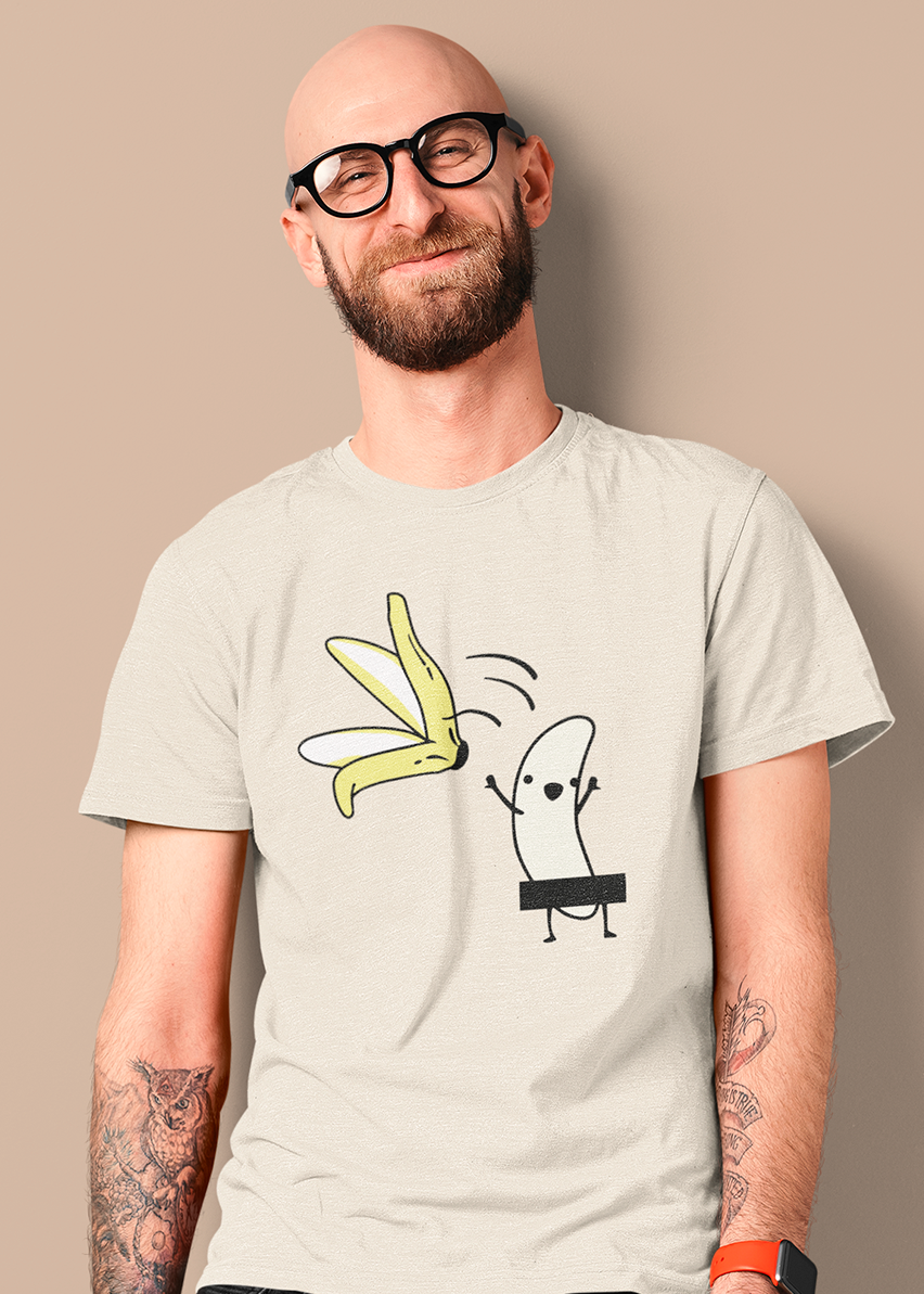 A man with a shaved head, beard, and black-rimmed glasses wearing a light beige graphic t-shirt. The shirt features a cartoon illustration of a banana peel falling on a small banana character.