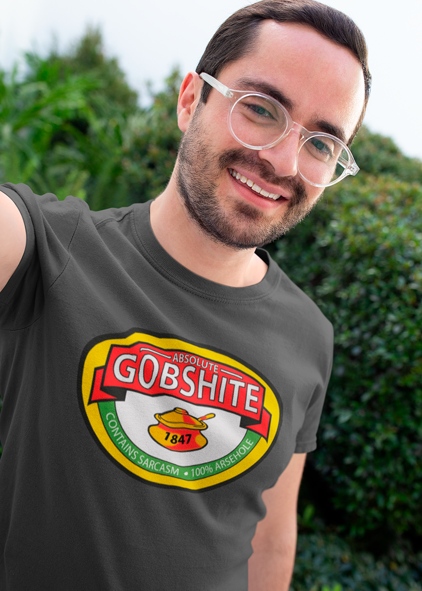 A man wearing a dark gray t-shirt with a parody graphic of the Gobshite brand logo, set against a blurred background of green foliage.