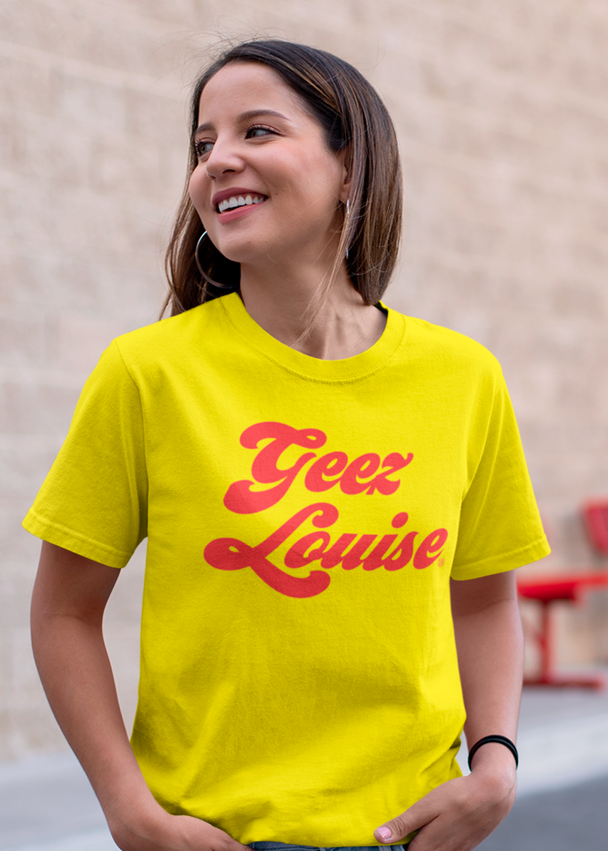 A woman wearing a bright yellow t-shirt with 'Geez Louise' printed in red cursive text across the chest, set against a blurred beige brick wall background.